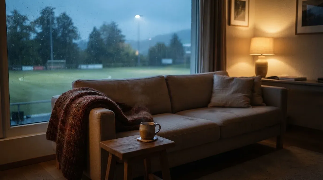 Ein Sofa mit Wolldecke steht vor einem grossen Fenster, draussen Regen und in der Ferne ein Fussballplatz.