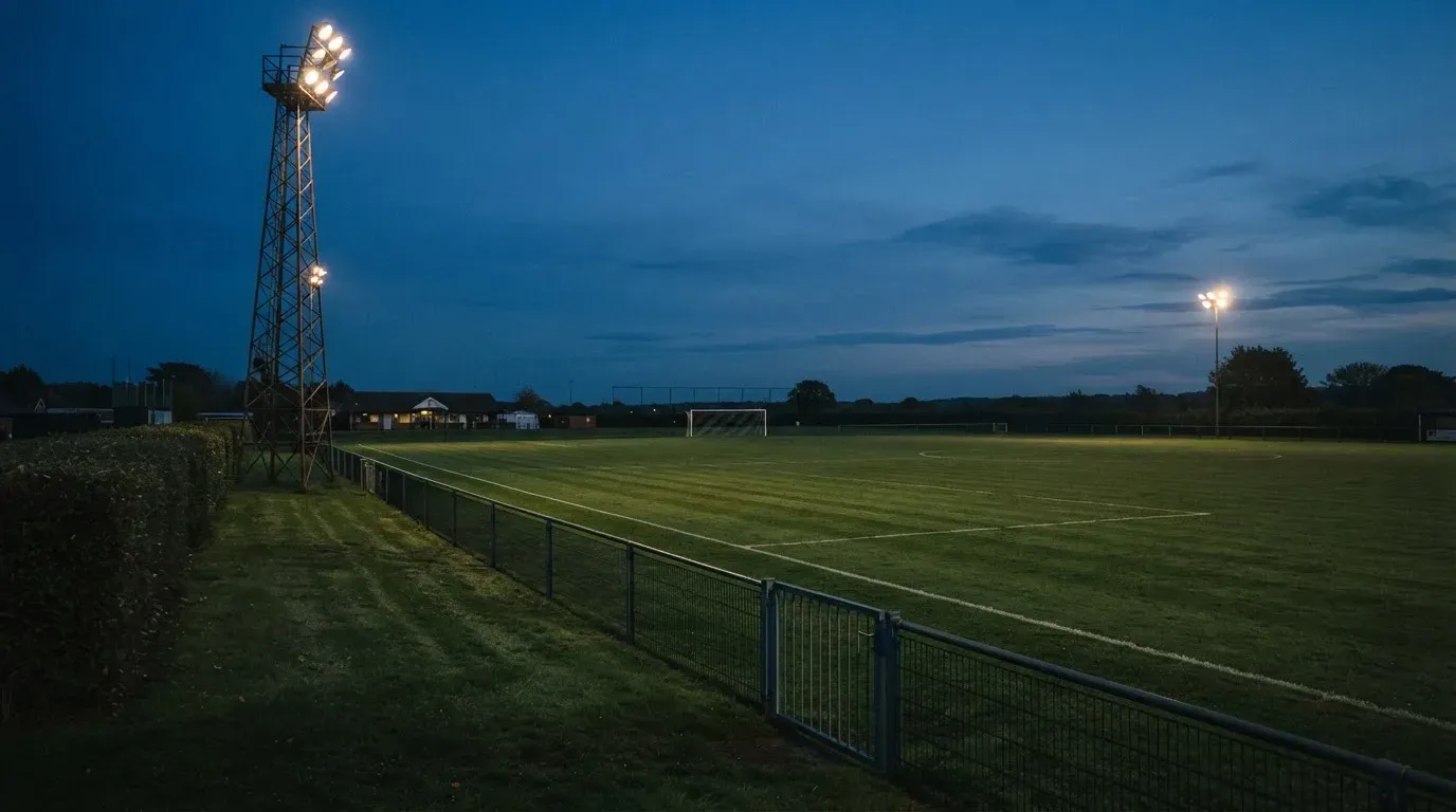 Ein Flutlichtmast ragt in den Abendhimmel, die Rasenfläche darunter liegt leer und dunkelgrün da.