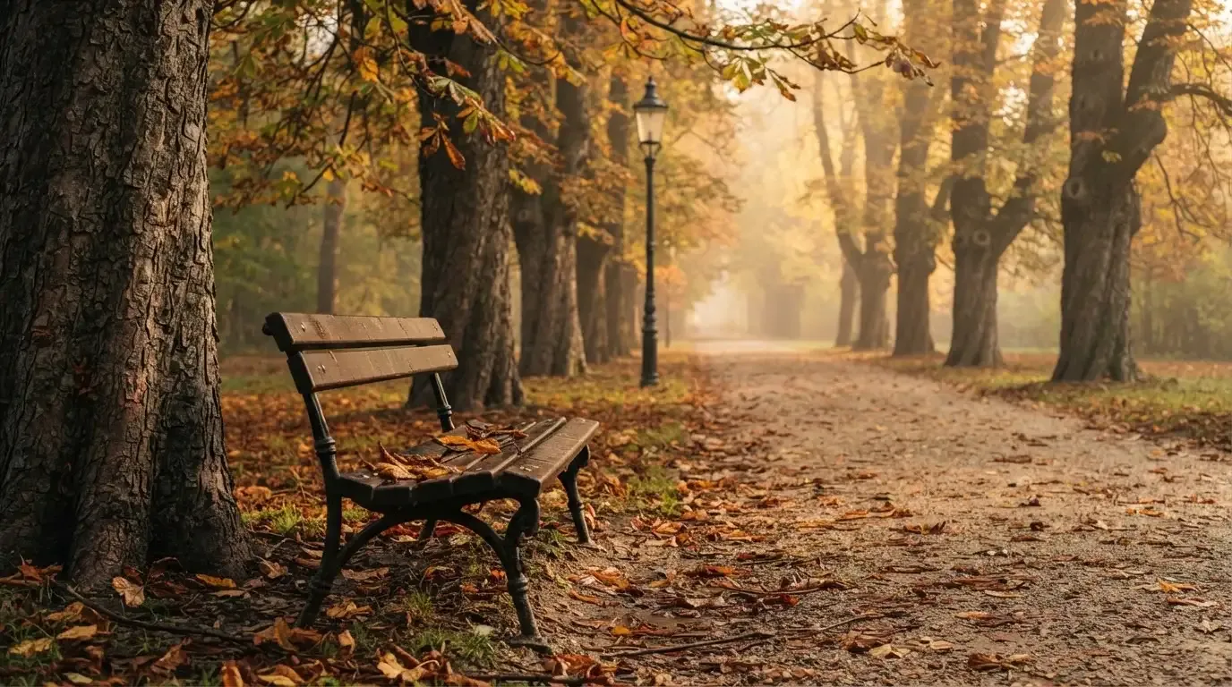 Eine Parkallee mit Kastanienbäumen und einer leeren Bank, Herbstlaub bedeckt den Kiesweg.