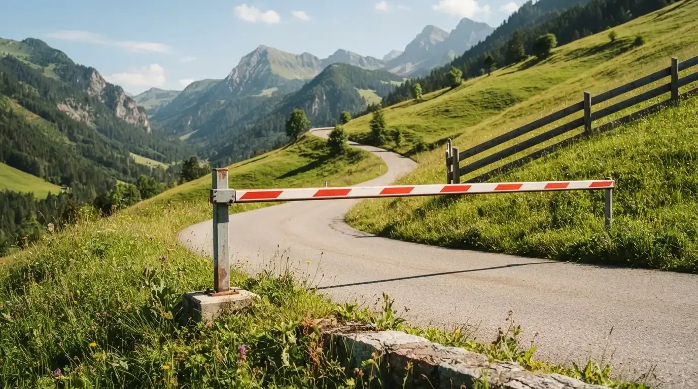 Ein Schlagbaum sperrt eine Landstrasse in den Bergen, dahinter windet sich der Weg zwischen grünen Hängen.