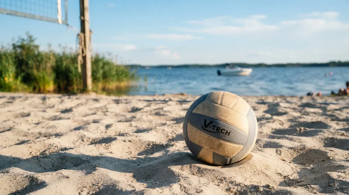 Ein Volleyball liegt im Sand eines Spielfelds am Seeufer, im Hintergrund gleitet ein Boot über das Wasser.