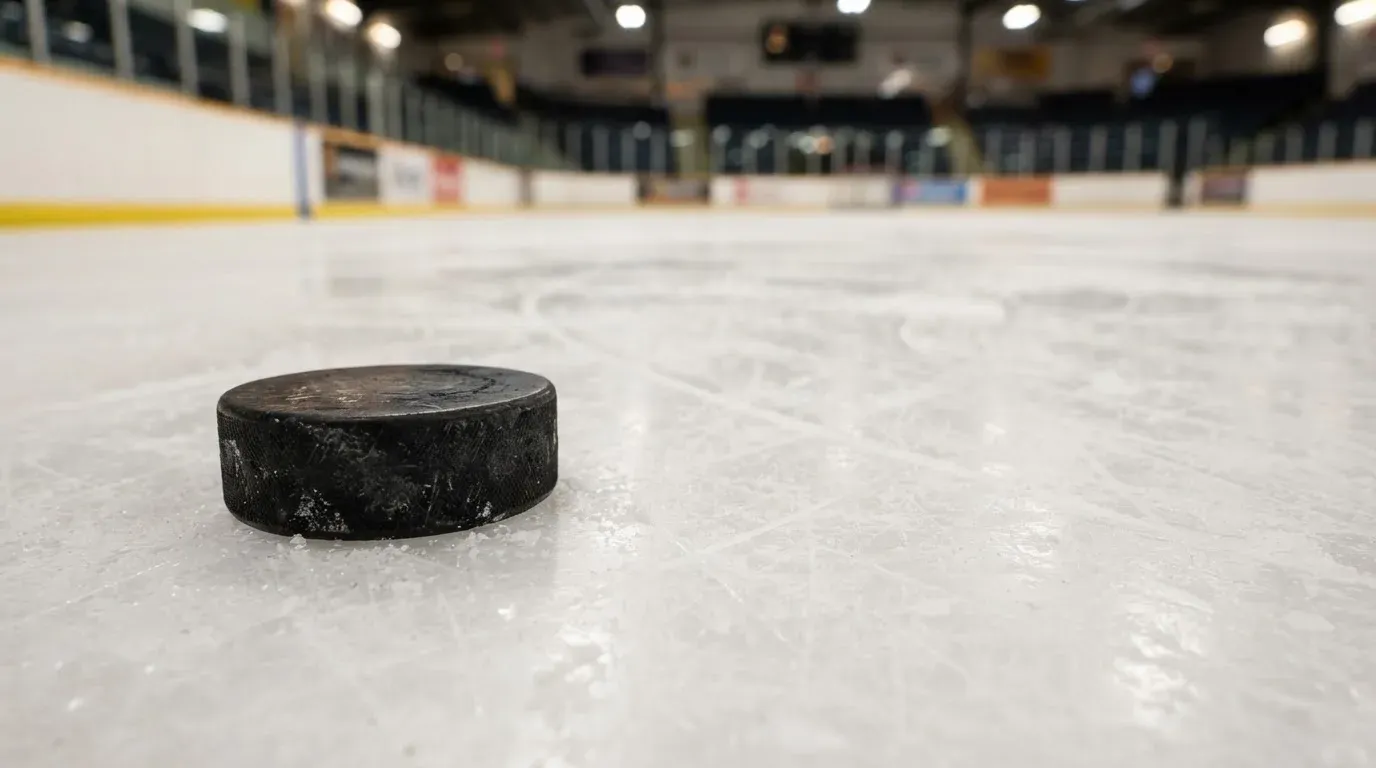 Ein Eishockeypuck liegt in Grossaufnahme auf der Eisfläche, feine Kratzer im Eis glänzen im hellen Hallenlicht.