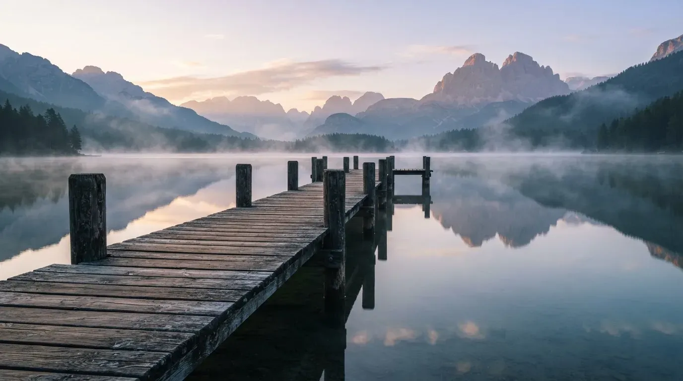 Ein Holzsteg führt über einen stillen Bergsee in den Alpen, über dem Wasser hängen bei Sonnenaufgang einzelne Nebelschwaden.