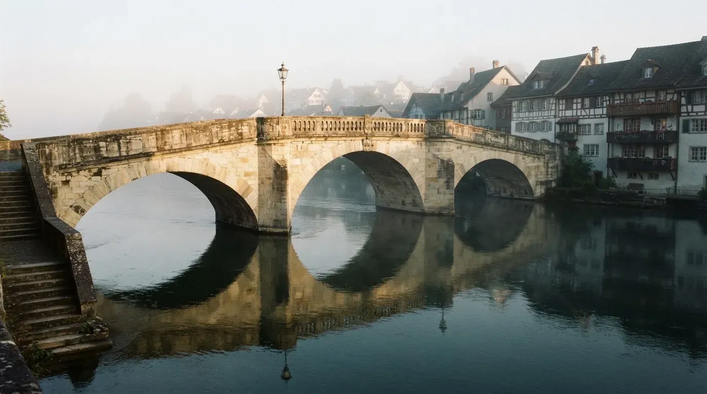 Eine steinerne Bogenbrücke überspannt am frühen Morgen einen Fluss in einer Altstadt, auf dem Wasser liegt dünner Nebel.