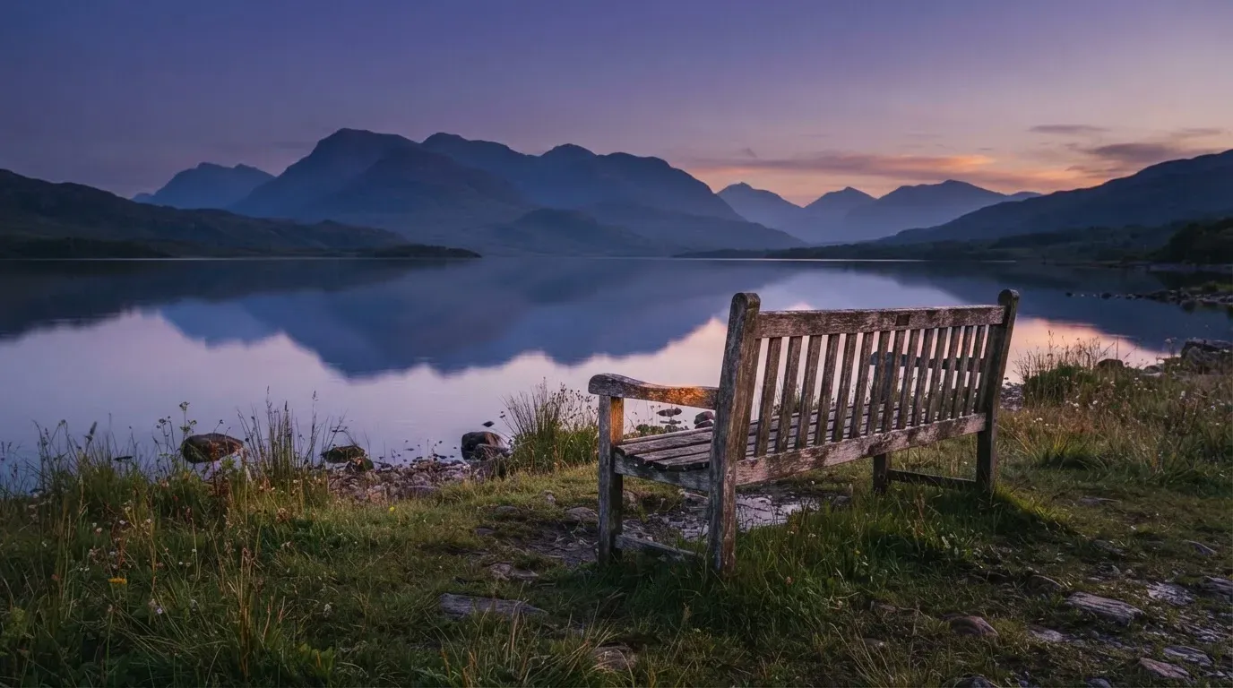 Eine Holzbank steht in der Dämmerung am Seeufer, im Hintergrund zeichnet sich ein Bergpanorama gegen den violetten Himmel ab.