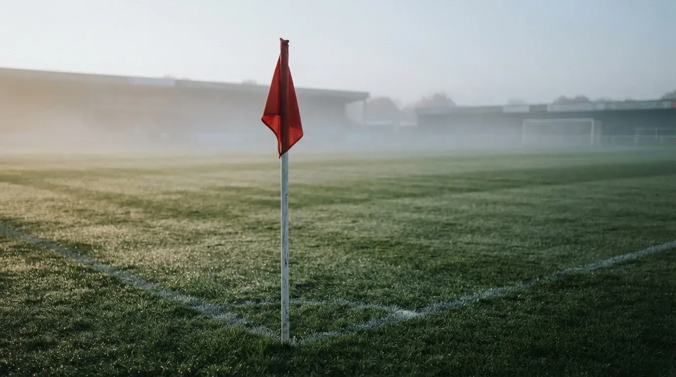 Eine Eckfahne steht am Morgen auf nassem Rasen, Tautropfen hängen im Gras und über dem Platz liegt leichter Dunst.