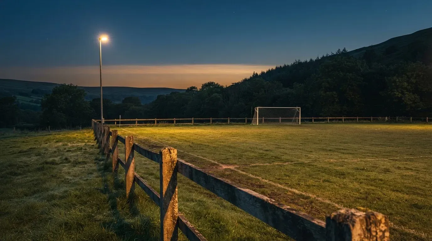 Ein kleiner Dorf-Fussballplatz liegt in der Dämmerung unter einem hohen Flutlichtmast, am Spielfeldrand steht ein Holzzaun.