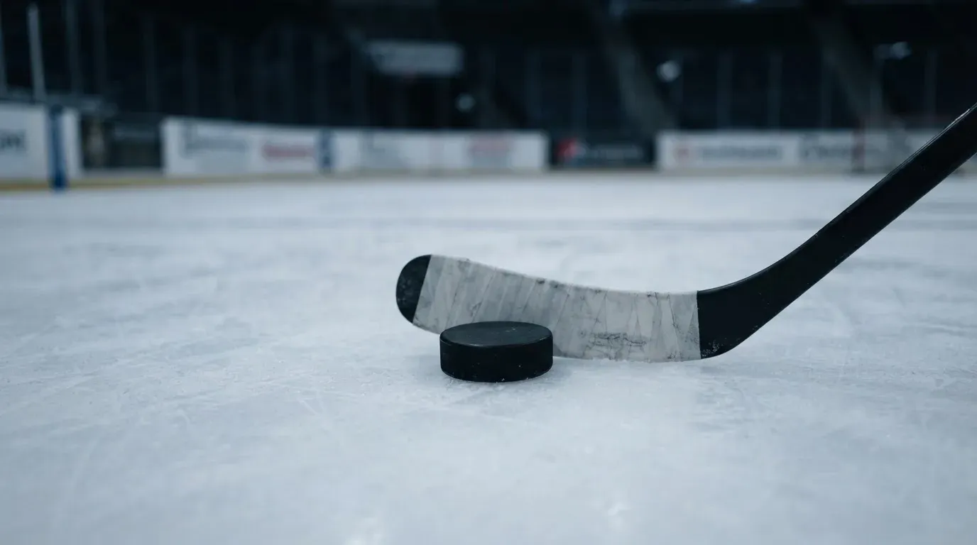 Ein Eishockeypuck liegt auf der Eisfläche neben dem Blatt eines Schlägers, die Bande dahinter ist im Dunkeln nur schwach zu erkennen.