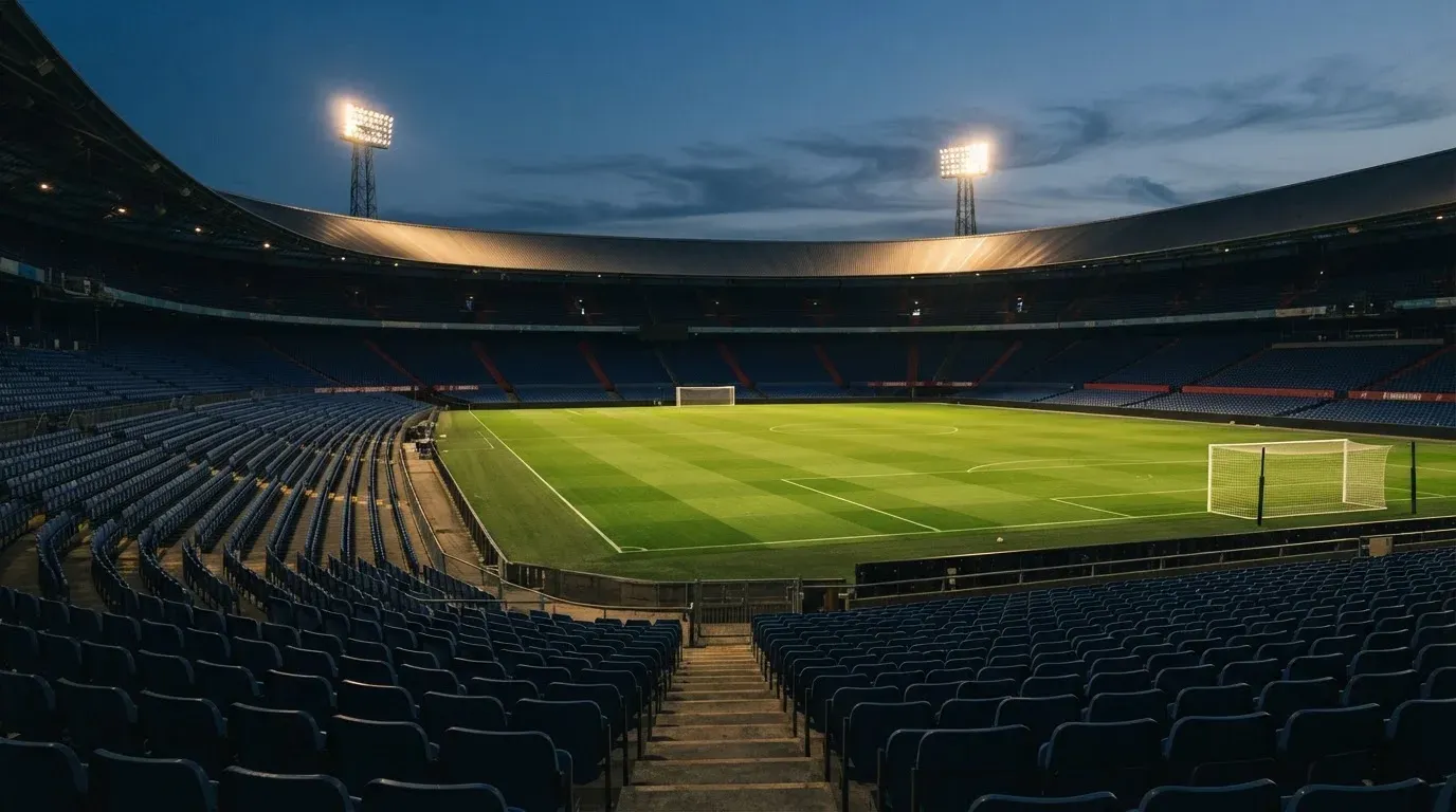 Leere Tribünenränge eines Fussballstadions in der Abenddämmerung, über dem Rasen leuchten die Flutlichter.