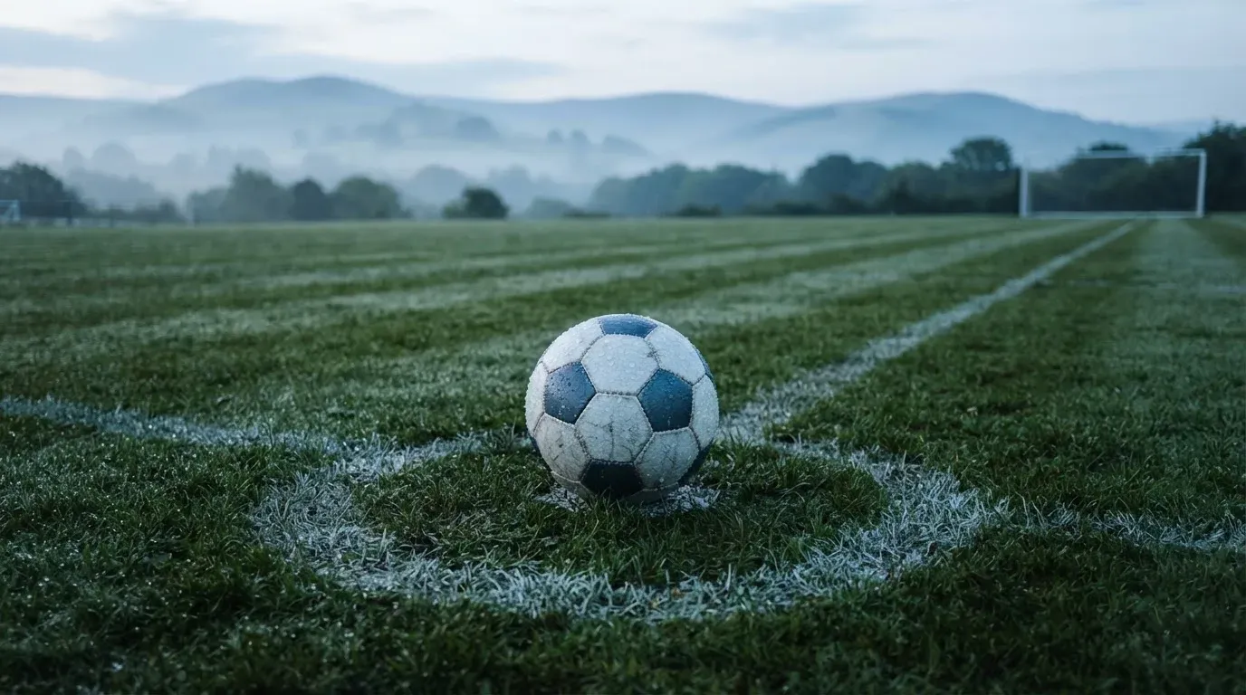 Ein Fussball liegt am frühen Morgen auf dem Anstosspunkt eines taunassen Rasens, im Hintergrund erheben sich Hügel im Dunst.