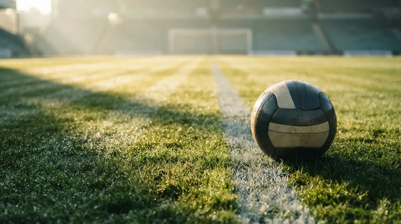 Ein Fussball ruht an der Seitenlinie im taufeuchten Gras, die ersten Sonnenstrahlen fallen schräg über den Platz.