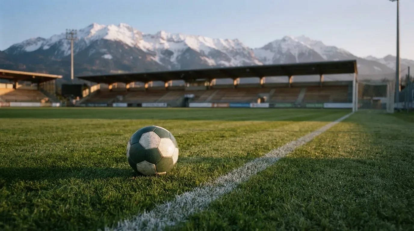 Ein Fussball liegt auf dem Rasen eines leeren Stadions am frühen Abend, dahinter erheben sich schneebedeckte Berge unter klarem Himmel.