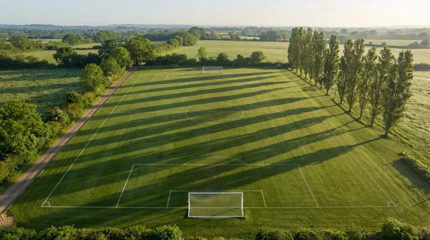 Ein leerer Fussballplatz mit weissen Linien von oben gesehen im Morgenlicht, am Rand stehen zwei Tore und eine Reihe Pappeln.