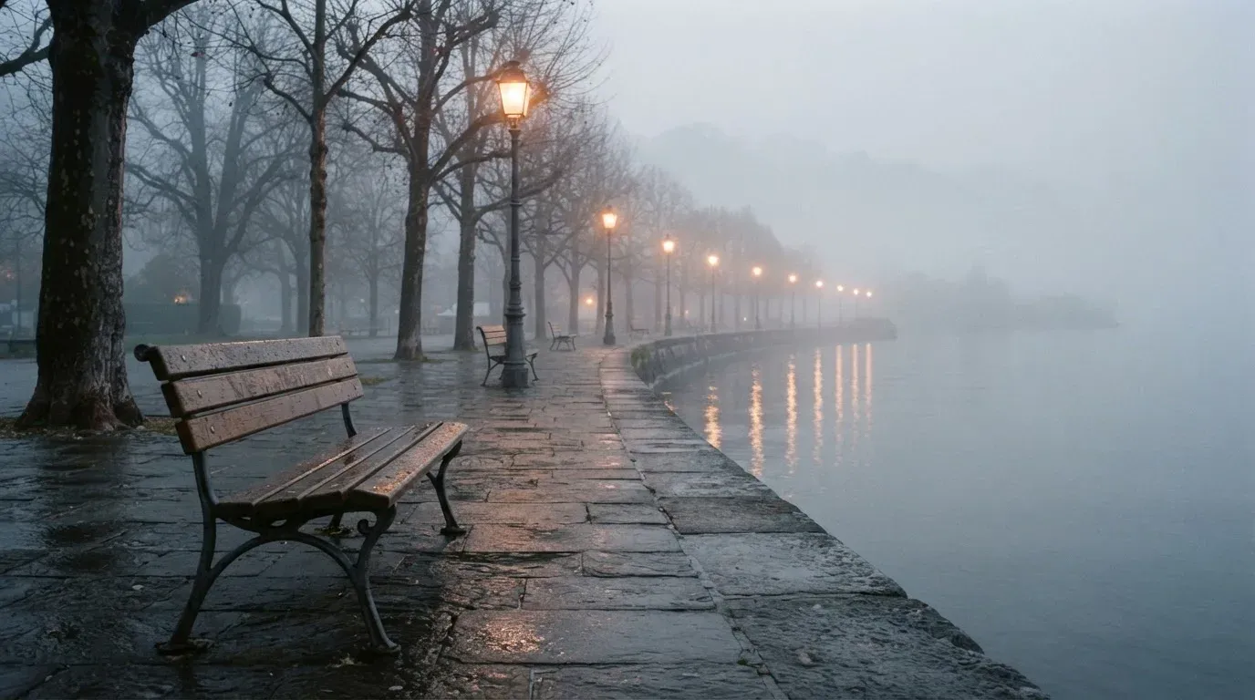 Eine Seepromenade mit leeren Bänken an einem nebligen Morgen, im Wasser spiegeln sich die Lichter der Uferlaternen.