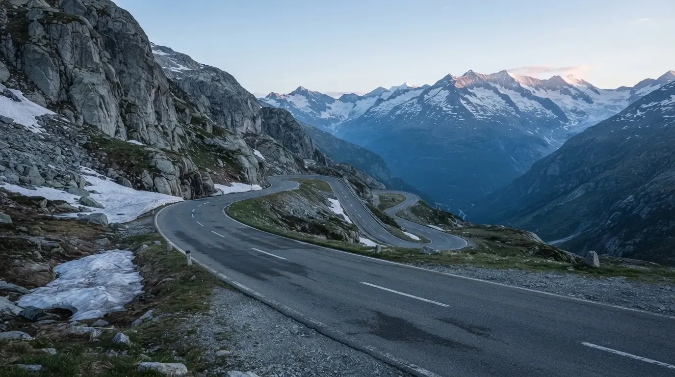 Ein Bergpass in den Schweizer Alpen mit leerer Strasse und Schneeresten am Rand im ersten Frühlicht.