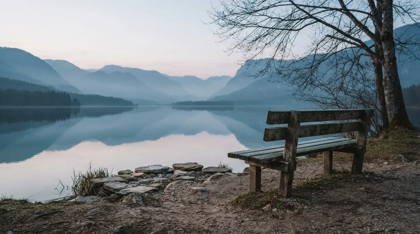 Eine Holzbank an einem Seeufer mit Blick auf eine Bergkette im Morgendunst, das Wasser ist spiegelglatt.