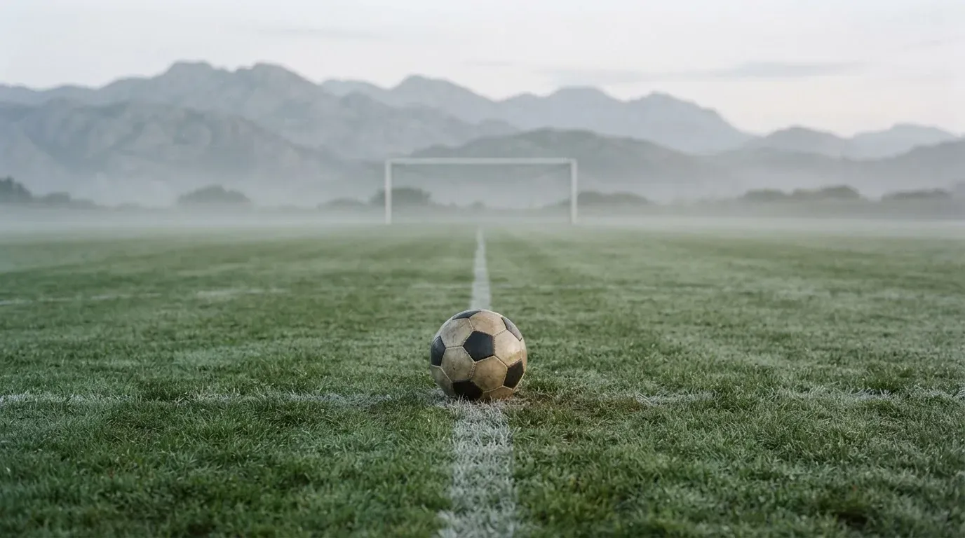 Ein einzelner Fussball auf dem Mittelpunkt eines leeren Rasens im Morgennebel, hinter dem Tor ragen Berge auf