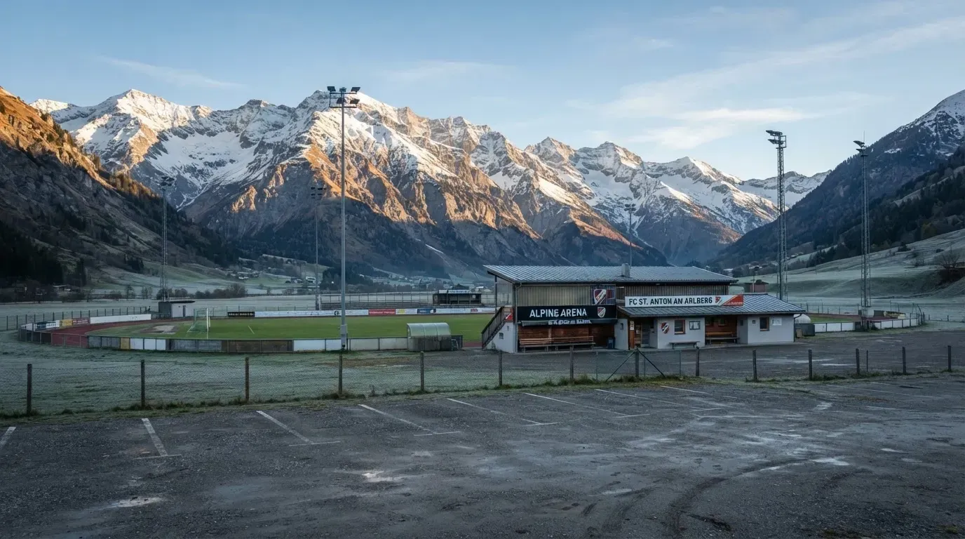 Schneebedeckte Berge ragen hinter einem kleinen Stadion in einem Alpental auf, im Vordergrund ein leerer Parkplatz am Morgen.