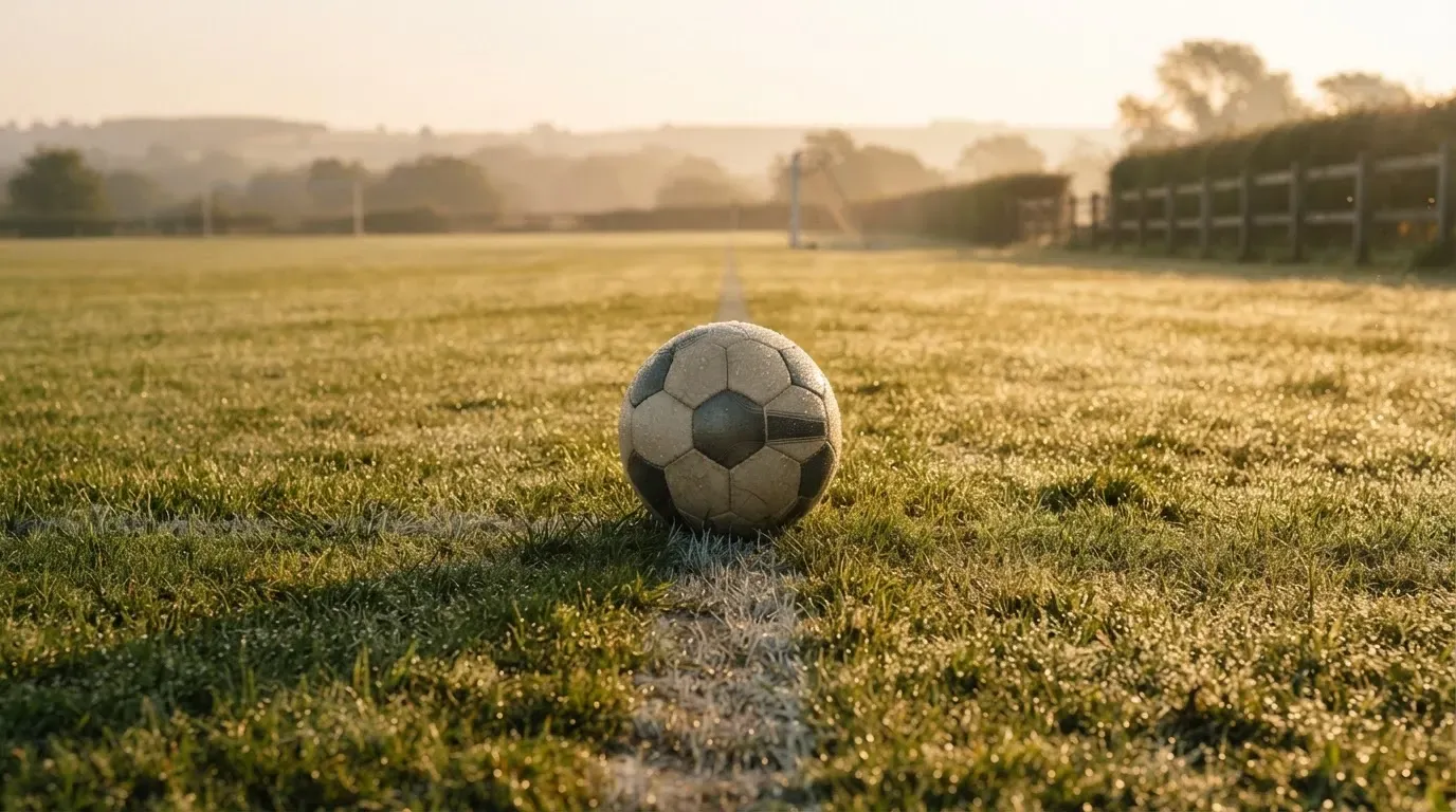 Ein Fussball liegt im Tau auf der Mittellinie eines Dorfplatzes bei Sonnenaufgang, der Rasen glitzert.