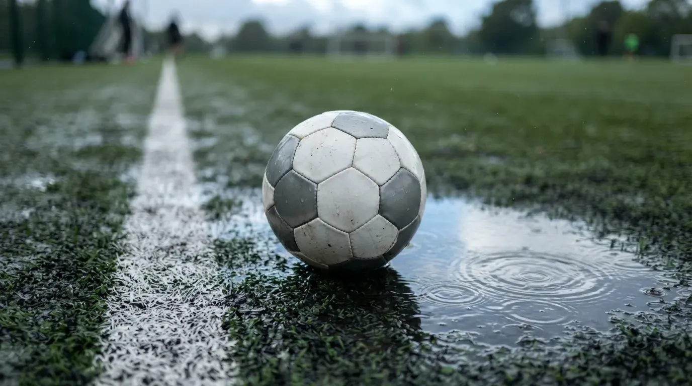 Ein Fussball liegt auf nassem Kunstrasen neben einer weissen Linie, Pfützen spiegeln den Himmel.