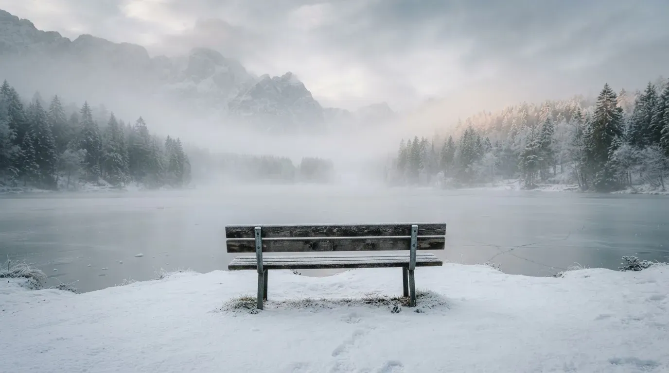 Ein zugefrorener Bergsee im Morgendunst, am Ufer steht eine einsame Holzbank zwischen verschneiten Tannen.