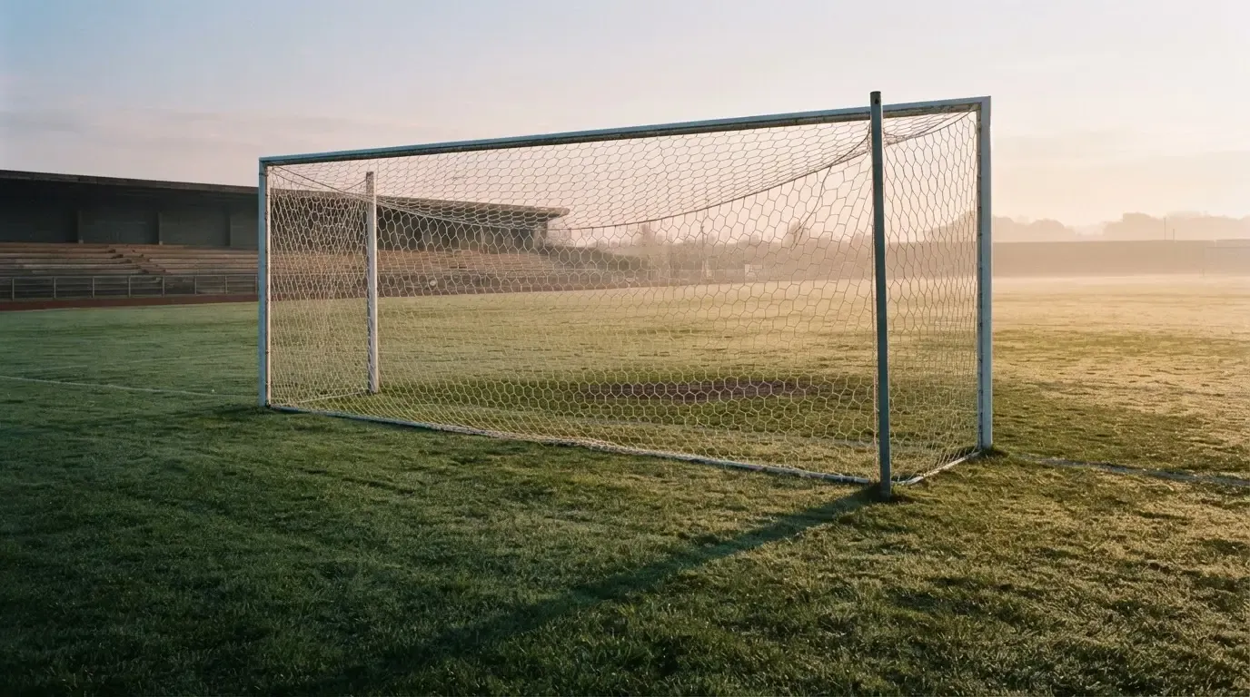 Ein Fussballtor mit Netz in der Totalen auf einem leeren Platz, Tau glänzt im Gras und die Morgensonne steht tief.