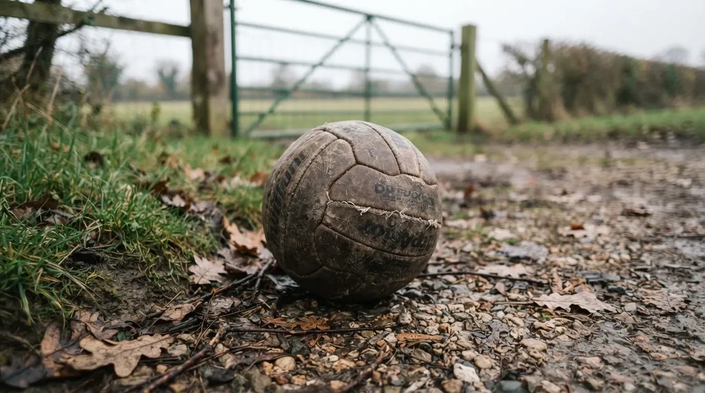 Ein abgenutzter Fussball liegt auf einem Kiesweg neben dem Eingang eines Vereinsplatzes, Laub liegt auf dem Boden.