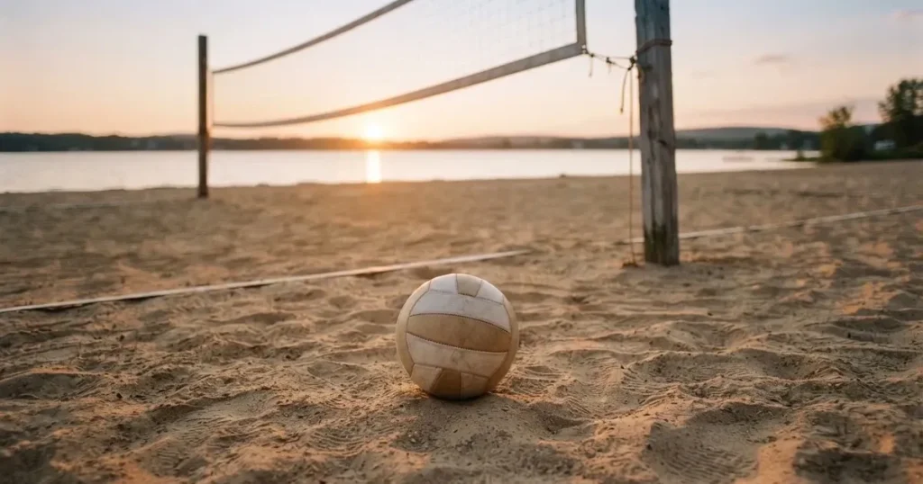 Ein Volleyball liegt im Sand eines Beachplatzes, zwischen zwei Pfosten ist ein Netz gespannt, tiefe Abendsonne wirft lange Schatten.