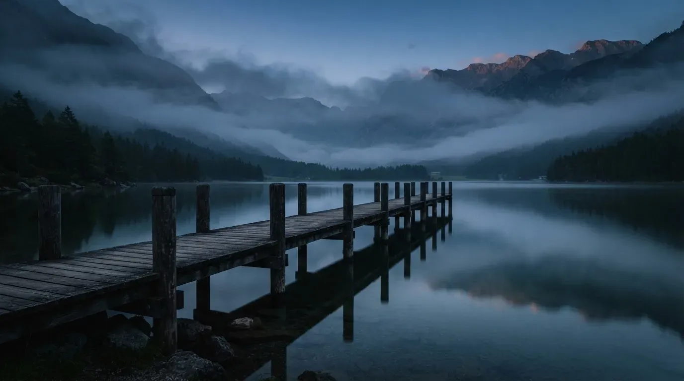 Ein Bergsee in der Dämmerung mit einem leeren Holzsteg, die Wasseroberfläche ist ruhig und Nebel zieht über die Hänge.