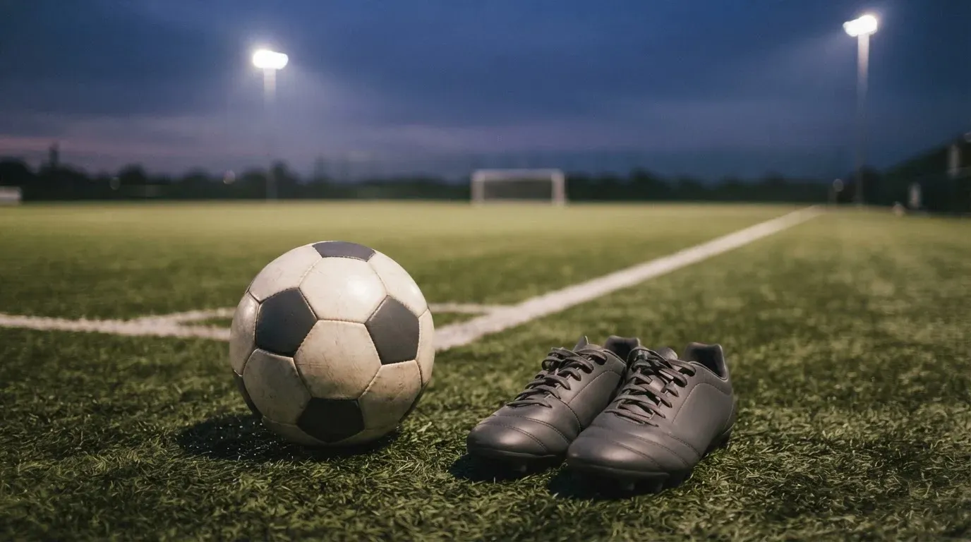 Ein Fussball und ein Paar Stollenschuhe stehen am Rand eines Kunstrasenplatzes, Flutlichtmasten ragen in den Abendhimmel.