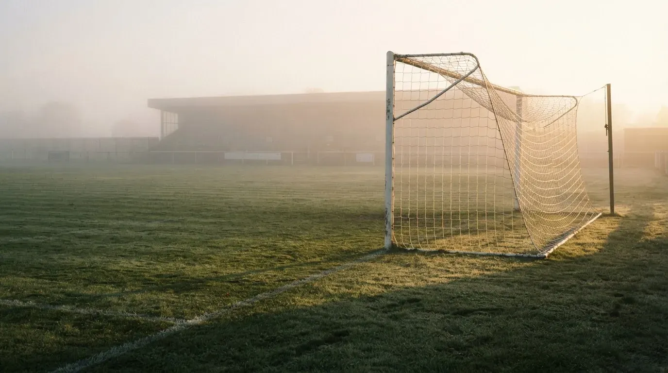 Ein Torpfosten mit Netz steht im Gegenlicht auf einem leeren Fussballplatz, über dem Rasen liegt Nebel.