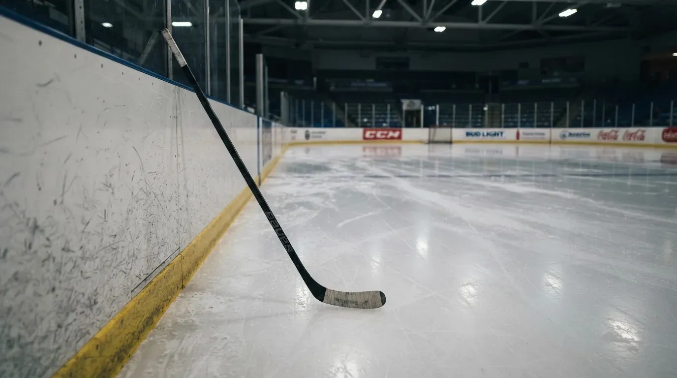 Ein Eishockeyschläger lehnt an der Bande einer leeren Halle, auf dem Eis spiegeln sich die Deckenlampen.