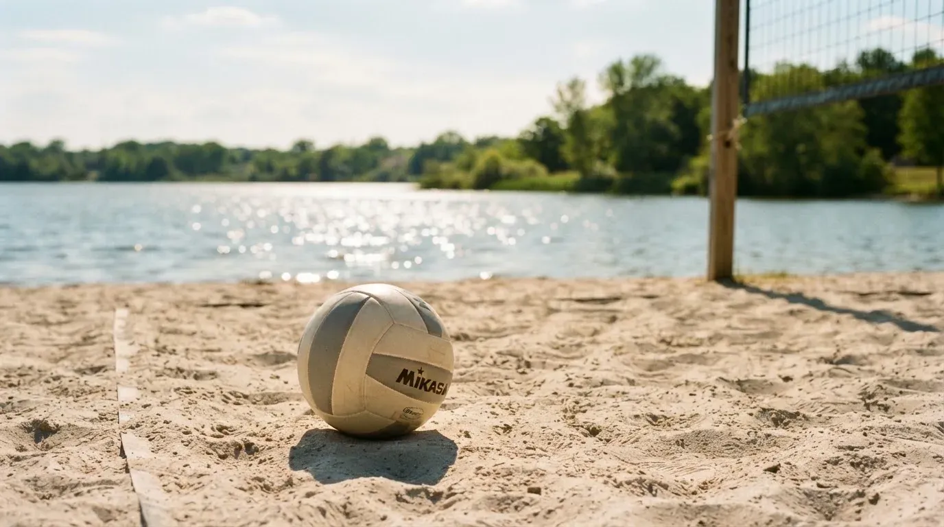 Ein Volleyball liegt auf dem Sand eines Strandfeldes am Seeufer, im Hintergrund glitzert das Wasser.