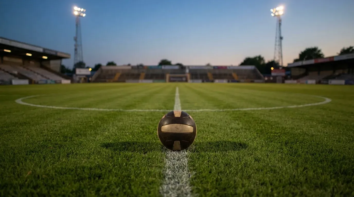 Ein Fussball liegt auf dem Mittelpunkt eines leeren Rasenplatzes, im Hintergrund ragen Flutlichtmasten in die Abenddämmerung