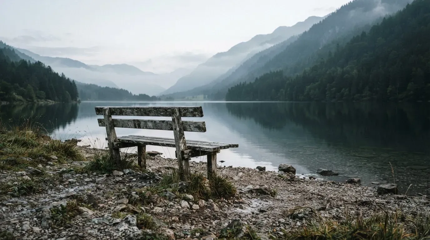Eine Holzbank am Ufer eines ruhigen Bergsees, am gegenüberliegenden Ufer steigen dunkle Hänge in den Frühnebel