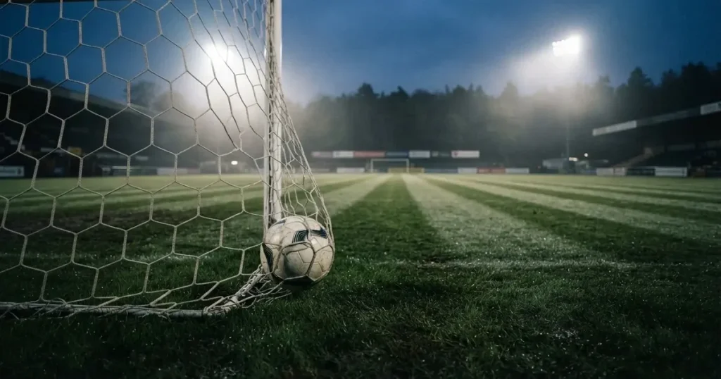 Ein Fussball liegt im Netz eines Tores, Flutlicht und kalte Abendluft liegen über dem leeren Platz