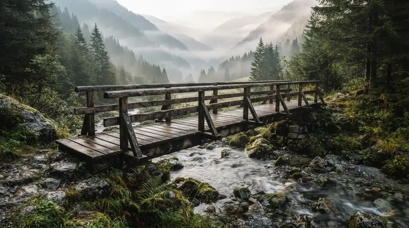 Eine alte Holzbrücke über einen Gebirgsbach, Moos auf den Steinen, Nebelschwaden hängen im Tal