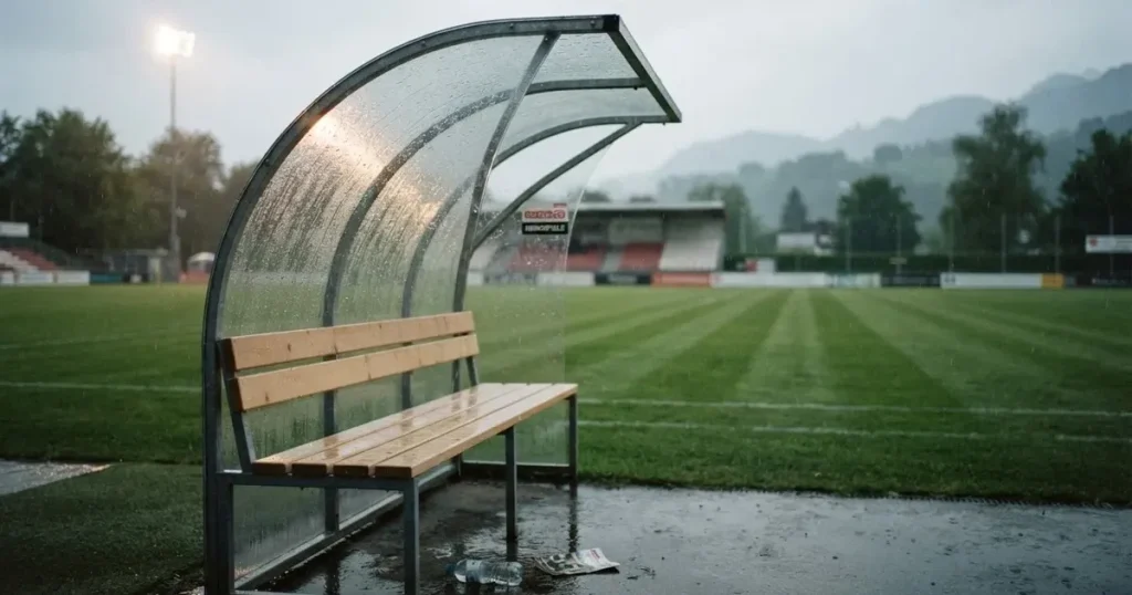 Eine leere Ersatzbank unter einem Plexiglasdach am Spielfeldrand, Regentropfen auf der Scheibe, dahinter ein Rasenplatz
