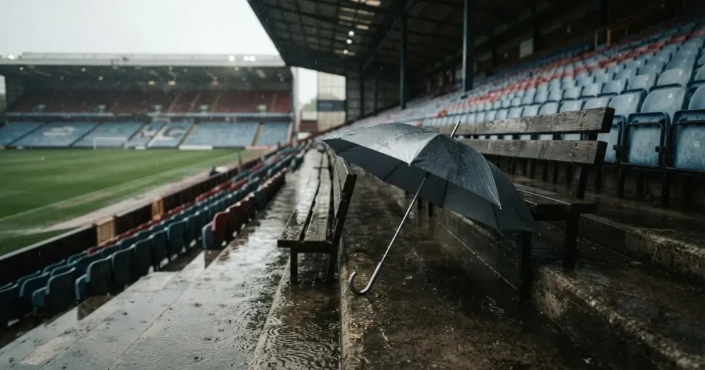 Ein offener Regenschirm lehnt an einer leeren Tribünenbank, Regen fällt auf die Stufen eines Stadions