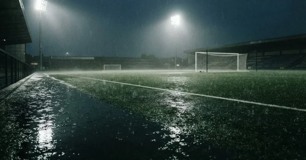 Ein Fussballplatz mit Kunstrasen im Regen, Pfützen spiegeln das Licht der Flutlichtmasten