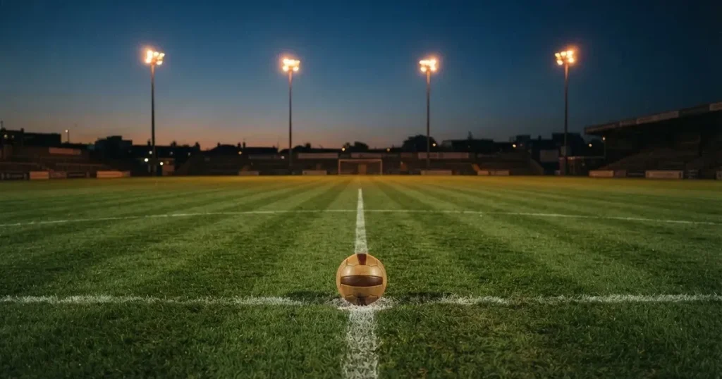 Ein Fussball liegt auf dem Anstosspunkt eines leeren Rasenplatzes in der Abenddämmerung, im Hintergrund leuchten Flutlichtmasten.