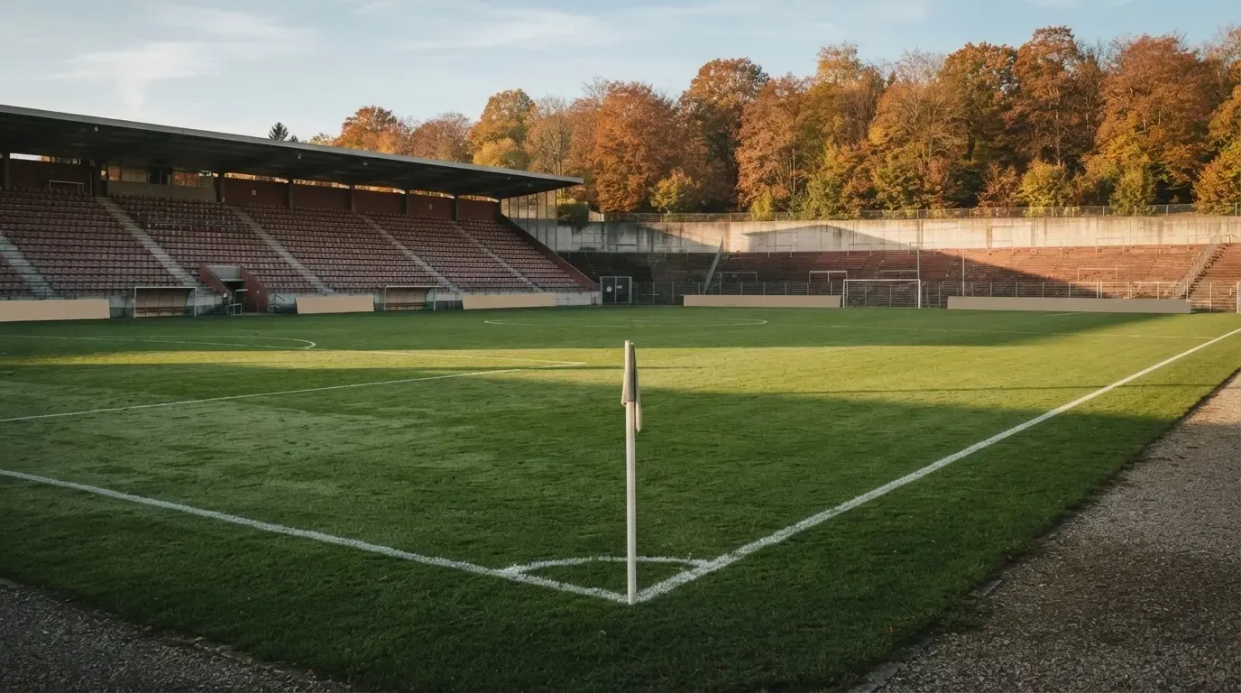 Leere Zuschauerränge eines kleinen Stadions im Herbstlicht, davor ein Rasenplatz mit frisch gezogenen Linien.