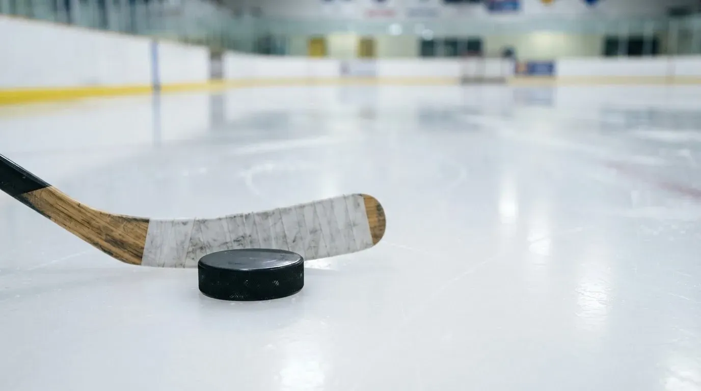 Ein Eishockeypuck und ein Schläger liegen auf einer glänzenden Eisfläche, im Hintergrund verschwimmen leere Banden.