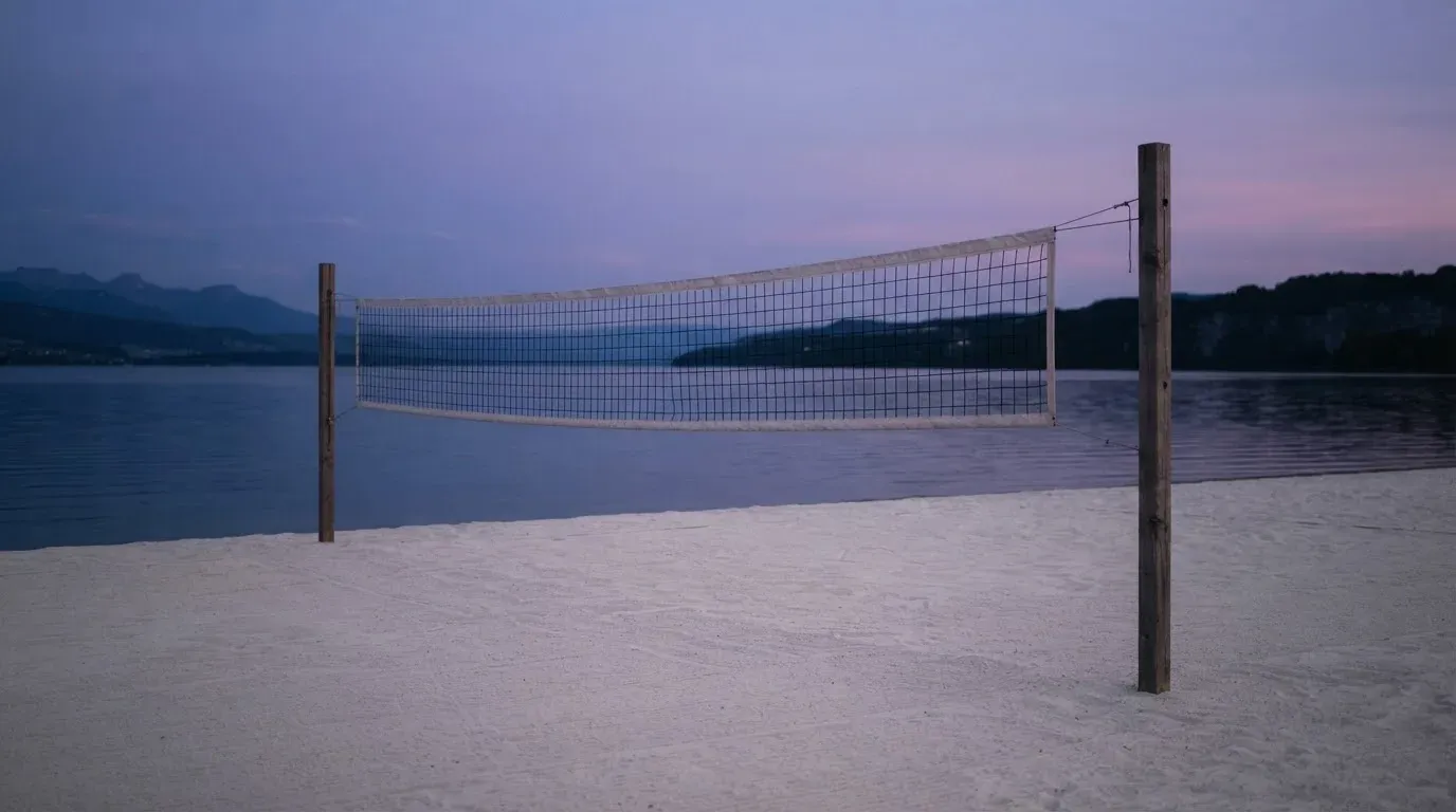 Ein Volleyballnetz steht an einem Seeufer in der Abenddämmerung, der Sand ist leer und das Wasser glatt.
