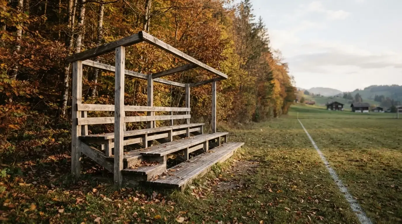 Eine leere Holztribüne steht am Waldrand neben einem kleinen Sportplatz im Herbstlicht.