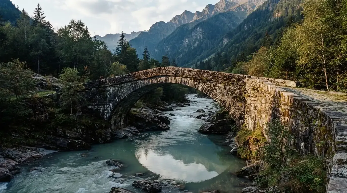 Eine alte Steinbrücke überspannt einen Gebirgsfluss, im Hintergrund steigen bewaldete Hänge an.