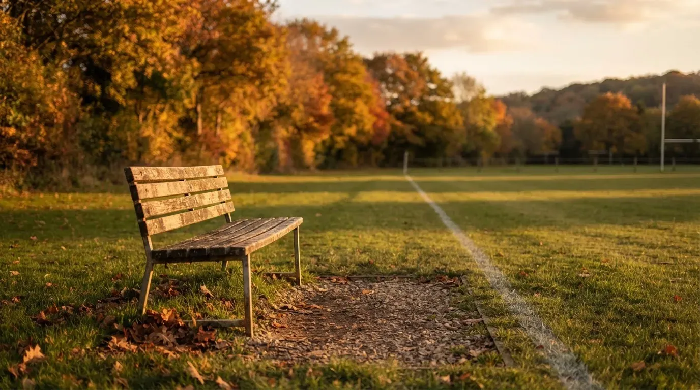 Eine hölzerne Bank steht an der Seitenlinie eines leeren Sportplatzes, dahinter Bäume im Herbstlicht.